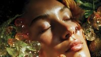Young African American woman with closed eyes surrounded by glowing crystals and green foliage in dreamy ethereal lighting creating magical atmosphere.