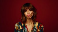 Young woman with bangs wearing colorful floral shirt making playful kiss expression against dark red background, fashion portrait with vibrant style and attitude.