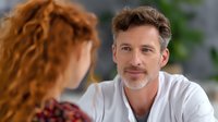 Mature Caucasian man with gray beard wearing white shirt smiling warmly during conversation with redhead woman in office setting, professional business meeting.