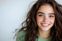 Young Hispanic woman with curly dark hair and bright genuine smile wearing green casual top against light background, radiating happiness and confidence.