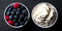 Fresh blueberries and raspberries in white ceramic bowl next to whipped cream dessert topping on dark slate background, overhead panoramic view.