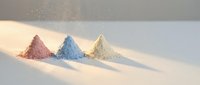 Three colorful powder piles in pink blue and beige creating artistic composition on white surface with soft lighting and scattered particles.