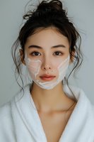 Young Asian woman wearing white bathrobe with facial cream mask treatment during skincare routine against light gray background, messy bun hairstyle and natural makeup look.
