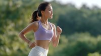 Young Asian woman in purple sports bra and white leggings jogging outdoors during morning workout against blurred green nature background. Active healthy lifestyle.