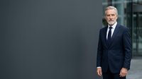 Mature Caucasian male executive in formal dark navy suit and tie standing against modern gray wall, suitable for corporate, business, and professional portraits.
