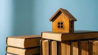 Small wooden house model sitting on stack of books against blue background representing real estate education and property investment knowledge.