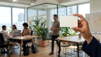 Cropped hand holding blank white card in modern office with diverse colleagues meeting in background, suitable for business communication and corporate branding projects.