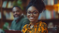 Young African American woman with glasses and natural hair smiling warmly in library setting, wearing vibrant yellow patterned blouse, with male student in background.
