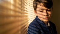Young Asian man in blue striped shirt by window with warm sunlight and shadow patterns from blinds creating dramatic lighting on face and wall.