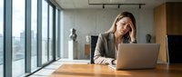 Stressed businesswoman with headache working on laptop in modern office. Professional female executive experiencing work pressure and fatigue.