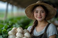 Young Asian female farmer in straw hat and overalls smiling while harvesting fresh white mushrooms in greenhouse, sustainable agriculture and organic farming concept.