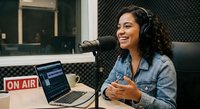 Young Hispanic woman radio host broadcasting live in professional studio with microphone, headphones and laptop showing audio software interface.