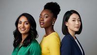 Three diverse professional women in colorful business attire standing confidently against gray background representing teamwork and corporate diversity.