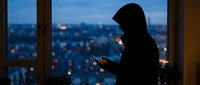Silhouette of hooded person using smartphone by window overlooking illuminated city at dusk. Anonymous figure browsing mobile device in urban apartment setting.