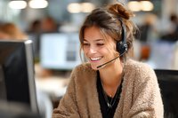Young female customer service representative with headset smiling while working at computer in modern call center office environment.