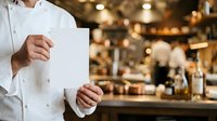 Chef in white uniform holding blank menu card in restaurant kitchen with blurred background, suitable for culinary branding and hospitality marketing.