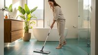 Woman in casual beige clothing cleaning modern bathroom floor with squeegee mop near white bathtub and potted plant for household cleaning and home maintenance.