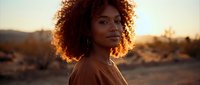 Young African American woman with curly hair in desert landscape during golden hour sunset, warm lighting creating natural glow for lifestyle photography.