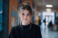 Young Caucasian woman with hair in messy bun wearing black sweater smiling at camera in blurred hallway background, conveying confidence and approachability.