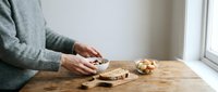 Woman in cozy sweater spreading jam on bread at wooden kitchen table near window creating warm homely breakfast scene.