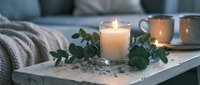 Cozy home interior with burning candle in glass jar, eucalyptus leaves and coffee cup on wooden tray. Warm hygge lifestyle scene for relaxation and comfort.