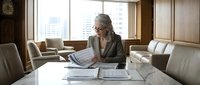 Senior woman with gray hair and glasses reviewing architectural blueprints and construction plans at conference table in modern office with city skyline view, suitable for business, architecture, and professional services.