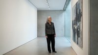 Professional blonde businesswoman in dark suit standing in modern minimalist art gallery with contemporary paintings and track lighting for corporate presentations.