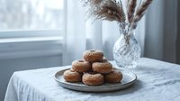 Freshly baked chocolate donuts arranged on ceramic plate beside crystal vase with pampas grass on marble table near window for cozy breakfast scene.
