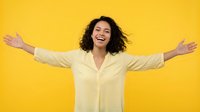 Woman with dark curly hair and medium skin tone wearing cream button-up shirt, arms spread wide in celebratory pose against bright yellow background, expressing joy and success for business, lifestyle, and motivational content.