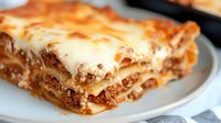 Homemade lasagna with layers of pasta, ground meat sauce, and melted cheese served on white plate, close-up view showing rich textures and layers.