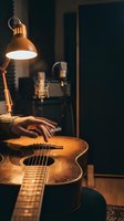 Musician's hand on acoustic guitar in warm home recording studio with professional microphone and desk lamp creating cozy atmosphere for music production.