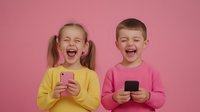 Two happy Caucasian children laughing while holding smartphones on pink background. Boy and girl enjoying mobile technology and digital entertainment.