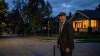 Senior Caucasian man with walking cane strolling through quiet suburban neighborhood at dusk with warm house lights glowing in background for lifestyle content.