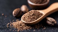 Ground nutmeg powder in wooden spoon with whole nutmegs scattered on dark background, macro food photography showing spice texture and aromatics.
