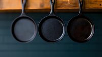 Cast iron skillets hanging on wooden rack against dark background, minimalist kitchen decor showcasing rustic cookware collection for food photography and culinary content.