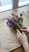Hands holding dried flower bouquet with purple statice, wheat, and thistle stems on kraft paper near bright window for floral design projects.