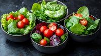 Fresh organic salad ingredients in black ceramic bowls with cherry tomatoes, spinach leaves, and lettuce on dark background. Healthy eating concept.