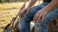 Senior man in denim jeans sitting on wooden fence in countryside field during golden hour sunset light for relaxation and outdoor lifestyle concepts.