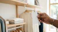Person with dark skin tone holding eyeglasses in a bright, minimalist bedroom with wooden shelving and folded clothing, suggesting optometry, vision care, or eyewear retail contexts.