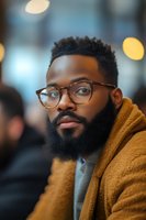 Young African American man with beard and glasses in brown jacket looking thoughtful in urban cafe setting with warm bokeh lights background.