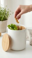 Hand dropping vegetable peel into white ceramic compost bin with bamboo lid on kitchen counter, sustainable zero waste lifestyle concept.