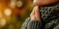 Close-up of hands in warm knitted sweater and scarf against blurred autumn background with bokeh lights, conveying cozy seasonal atmosphere.