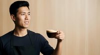 Asian male in his twenties wearing black casual clothing, holding a glass of dark beverage against warm wooden background, smiling while looking to the side. Suitable for lifestyle, beverage, and hospitality marketing.