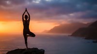 Woman practicing yoga tree pose on rocky cliff overlooking ocean at sunset with dramatic colorful sky and mountain silhouettes for wellness meditation.