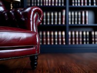 Luxurious burgundy leather armchair with ornate details against law library background with rows of books. Classic professional interior setting on wooden floor.