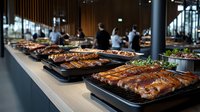 Grilled ribs and meat dishes on buffet table in modern restaurant interior with dark columns and blurred people in background, catering service setup.