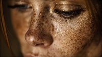 Close-up portrait of young woman with freckles and closed eyes, face partially visible with golden light highlighting natural skin texture.