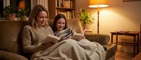 Mother and daughter reading book together on cozy sofa under warm lamp light in comfortable living room. Family bonding time at home during evening.