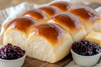 Fresh baked Japanese milk bread rolls with golden brown crust served on wooden board with sweet berry jam in small ceramic bowls, close up view.