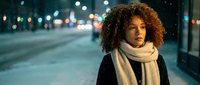 Young African American woman with curly hair wearing white scarf on snowy city street at night with bokeh lights creating warm urban winter atmosphere.